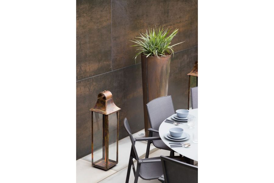 Tall empty copper and glass lantern and tall pot stand against wall faced with Steel Dark DesignClad panels, behind dining set.