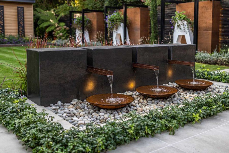 Water flows from low wall faced with Steel Dark external cladding into shallow bowls set on pebbles edged with low-growing shrubs.