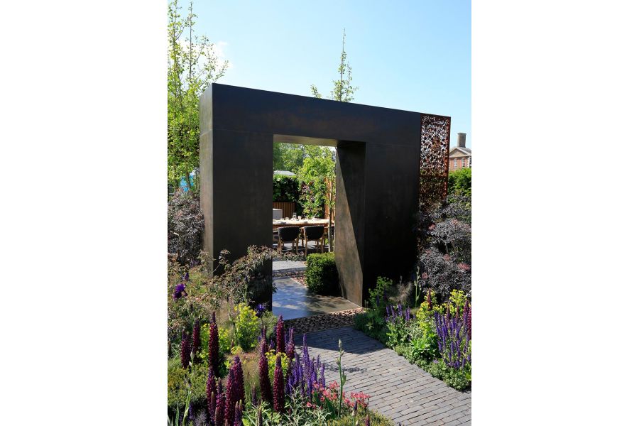 Chunky square arch faced with Steel Dark external cladding straddles path leading between planted beds to seating area.