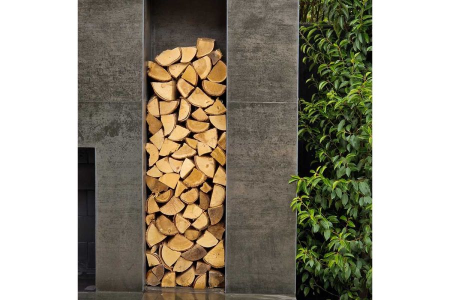 Split logs piled in tall niche between walls faced with Steel Dark external cladding. Bush to right.  Design by Sapphire Externals.