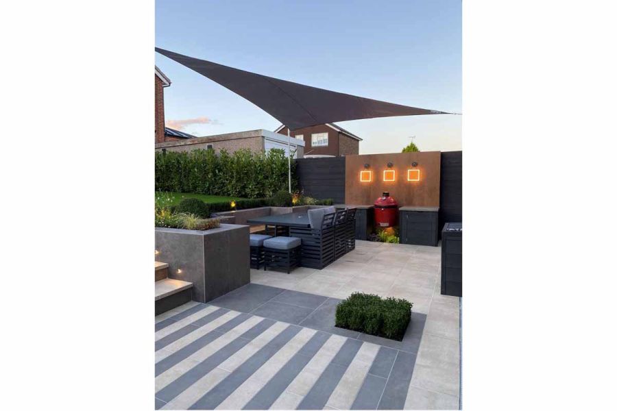 Alternate steel-grey and cream porcelain patio tiles lead to steps to lawn level, from patio with outdoor kitchen and shade sail. 