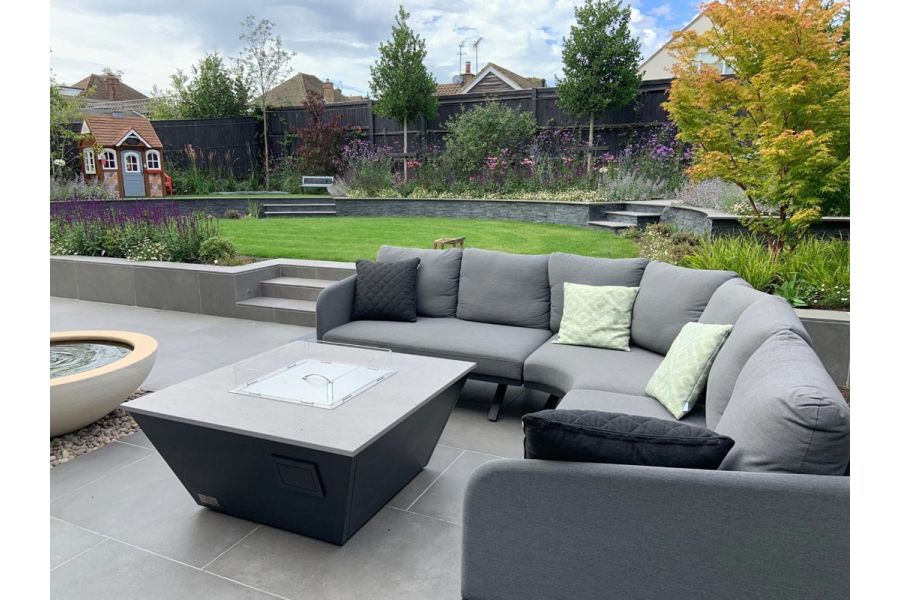 Large outdoor curved sofa around a firepit sitting on a Steel Grey porcelain patio and in front of low retaining wall leading to a raised lawn.
