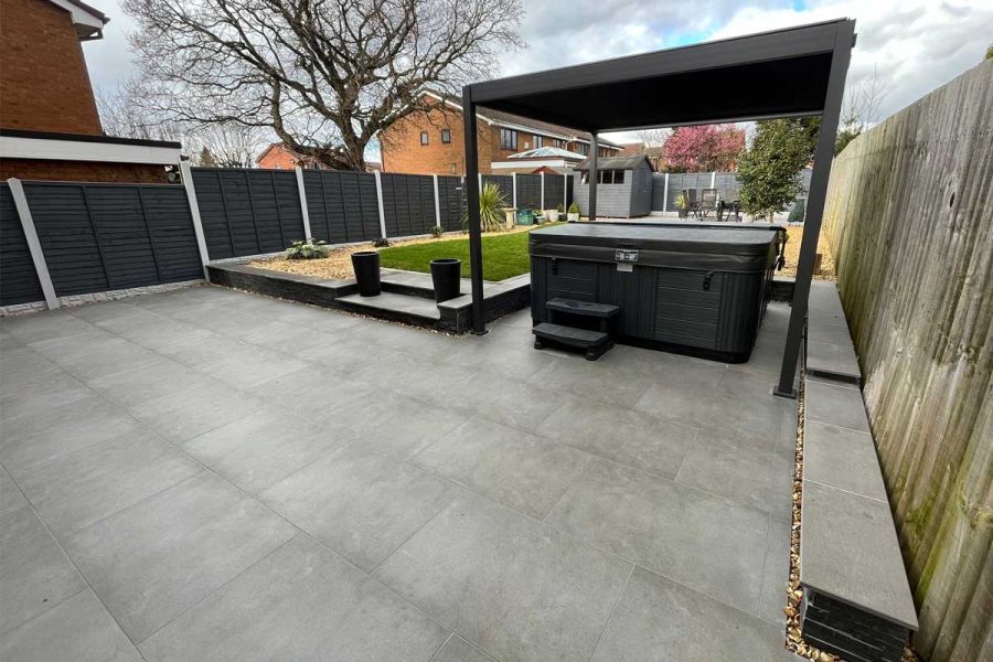 600x600 sized Steel Grey porcelain pavers leading towards a raised lawn and hot tub underneath a metal pergola.