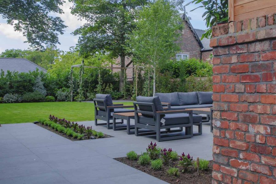 View of garden with Steel Grey porcelain paving slabs, with luxury garden furniture, to fencing lined with shrubs.