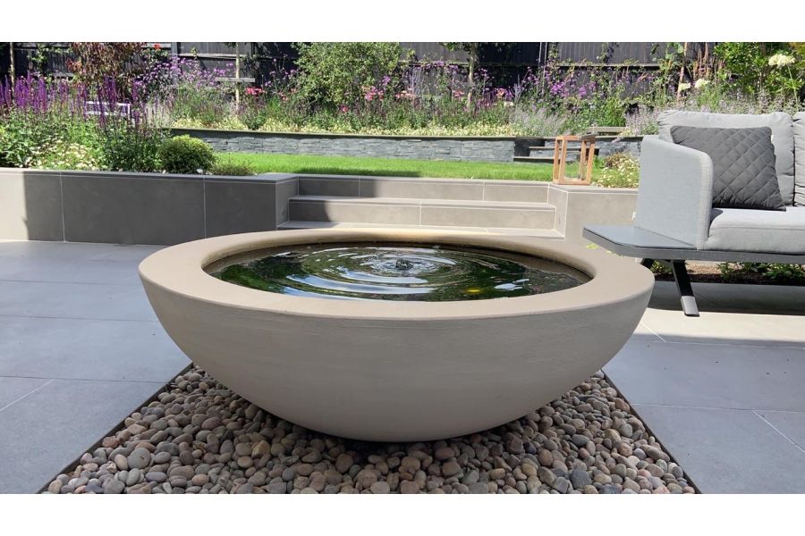 Large stone bowl water feature sitting on a bed of smooth pebbles and surrounded on all sides by Steel Grey porcelain paving.