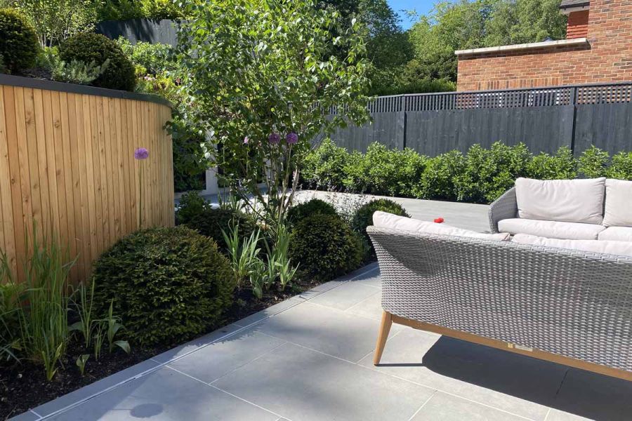 Cosy back garden paved in Steel Grey porcelain with flower beds planted with box hedging and a set of wicker garden sofas.
