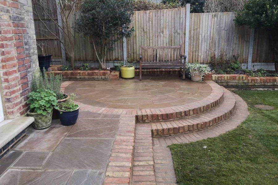 Narrow Raj Green Sandstone patio with Sandstone Paving Circle at corner of house. 3 straight and curved 2 brick steps up from lawn.