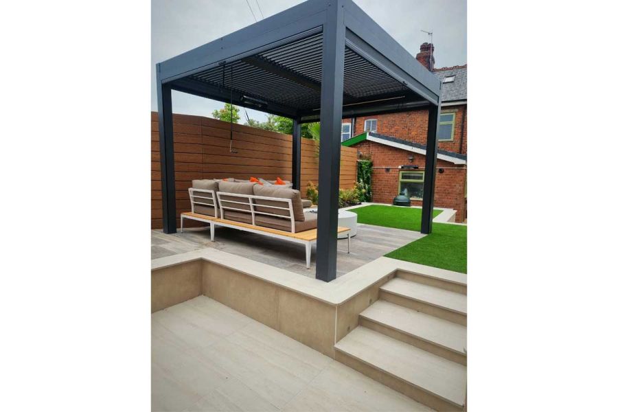 Faro Porcelain paving and steps up to metal pergola over modular corner sofa in raised area of garden with lawn, beds and fencing.