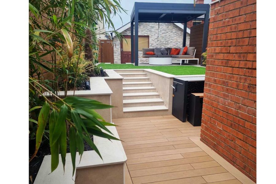 Mocha DesignBoard WPC decking between brick wall and bamboo in raised bed, with 8 porcelain steps up to lawned area with pergola. 