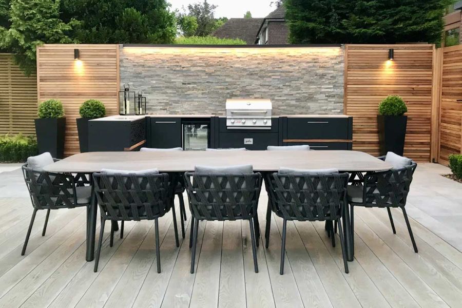 Outdoor kitchen with stone-clad back wall on edge of dining area laid with Smoked Oak Millboard decking by Landscape Design Studio.