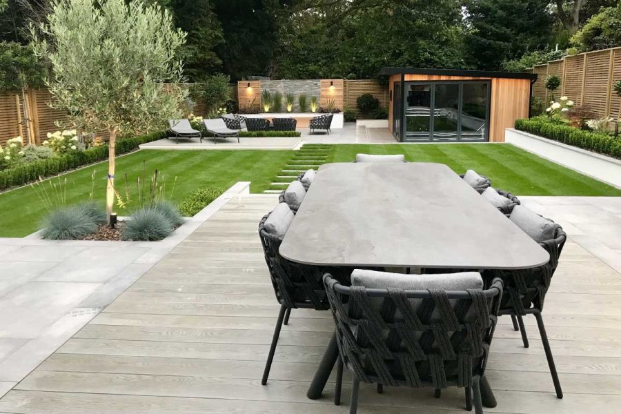 8-seater table on Millboard Smoked Oak decking boards laid flush with paving slabs in garden with lawn, studio and seating area.