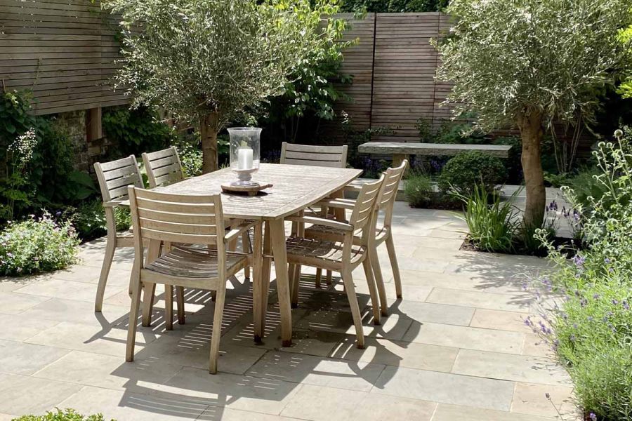 Wooden dining set on Heath sawn sandstone paving in tall-fenced garden with olive trees and planted borders. 