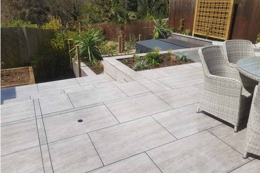 Travertine Dark porcelain paving patio by Living Landscapes, with matching grey rattan furniture. Steps descend past raised beds.