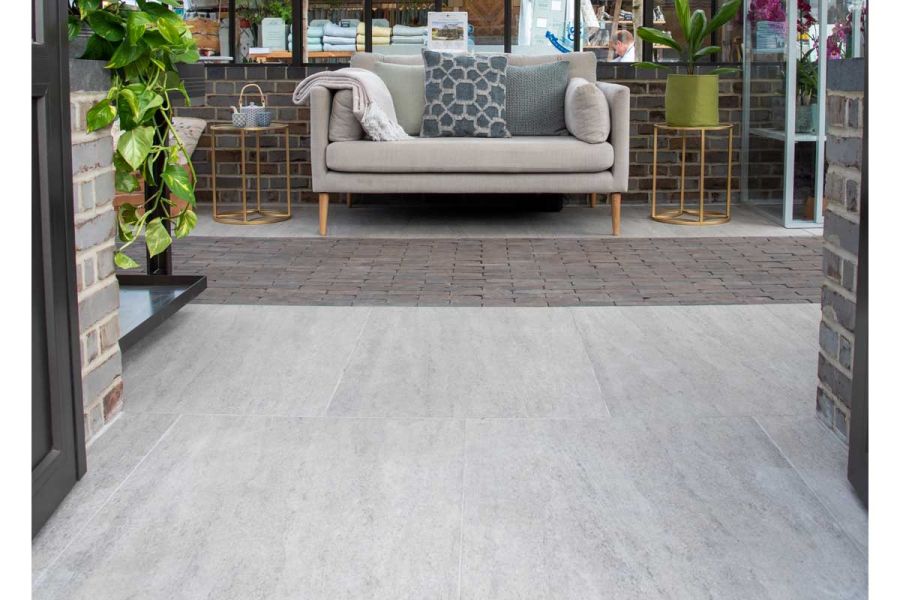 Sofa sits on Travertine Dark Porcelain tiles against wall in half-bricked greenhouse, tables on either side. Design by Llevelo.