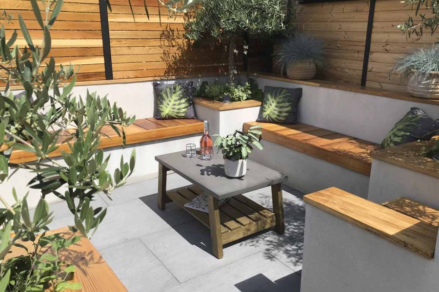 Corner of seating area. White rendered, wooden-coped walls, benches and raised square beds surround trendy black porcelain paving.