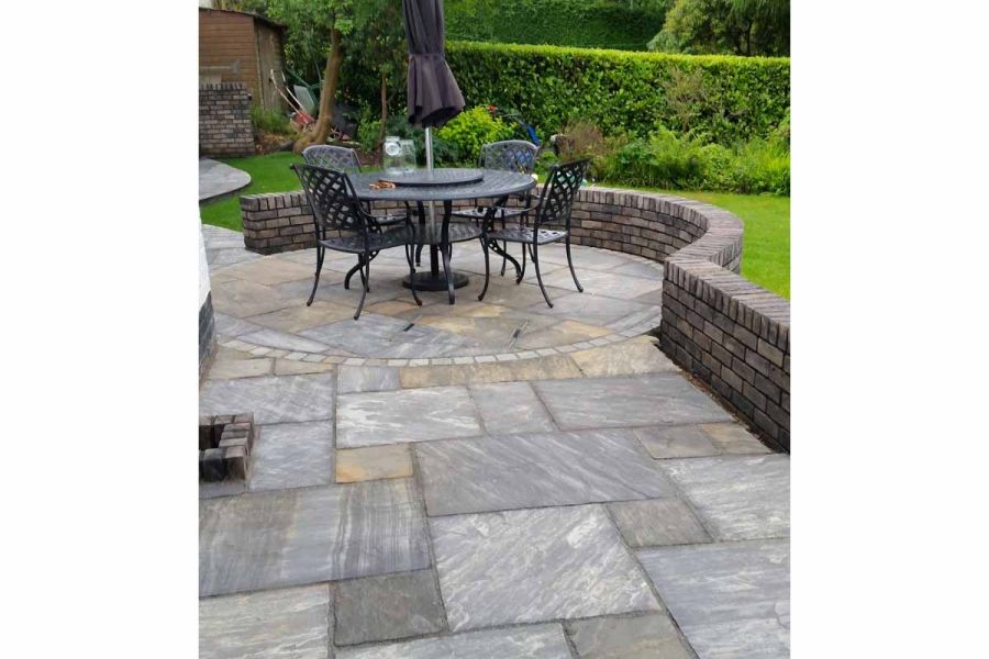 Paving circle forms part of back garden patio of Tumbled Black sandstone paving, separated from hedged lawn by low brick wall. 