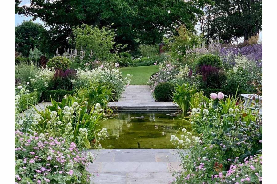 Large square pond divides Tumbled Black Indian sandstone path leading to lawn beyond between planted borders. 