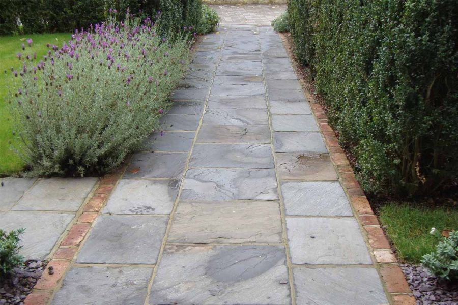 Straight front garden path of Tumbled Black sandstone paving with brick paver edge leads between planted border to front gate.