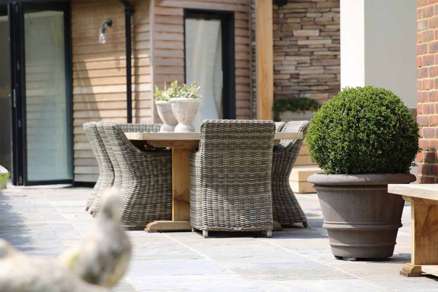 Topiary ball in pot sits on sunny Black tumbled Indian sandstone paving with outdoor furniture with french doors in background.