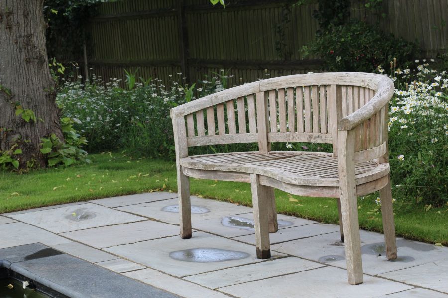 Curved-back wooden bench stands on tumbled grey Indian sandstone paving, with mown grass, flower border and mature tree behind.