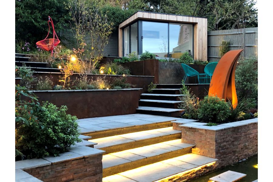 Studio at top of sloping garden terraced with brick-faced raised beds and ascended by Kandla Grey tumbled sandstone steps. 