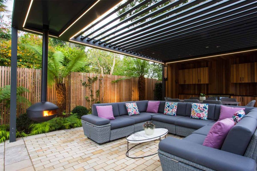 Large modular sofa on Tumbled Mint cobble setts, under louvred metal pergola with hanging woodburner. Fence and tree ferns behind.