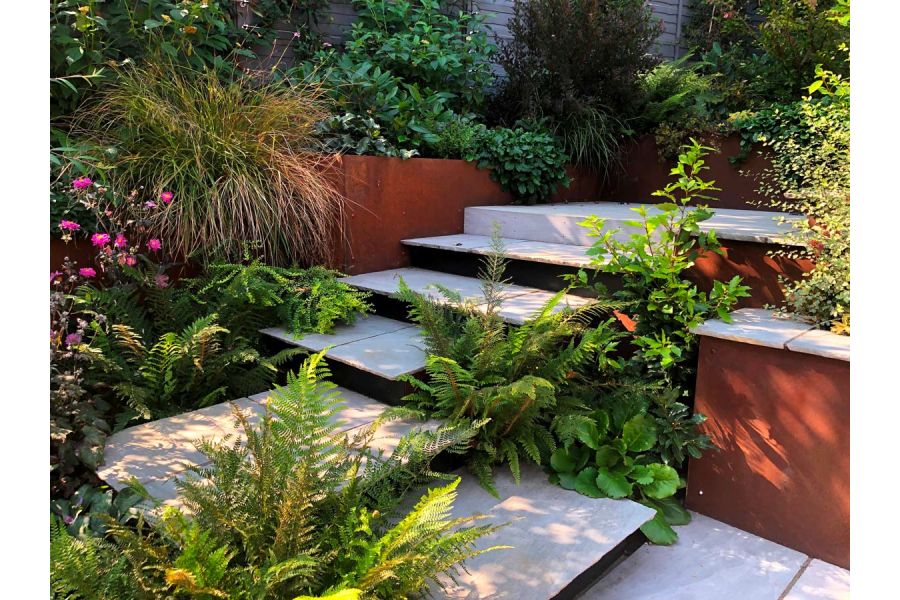 Steps of tumbled Kandla grey garden paving slabs turn right angle and rise to paved area surrounded by lush planting.