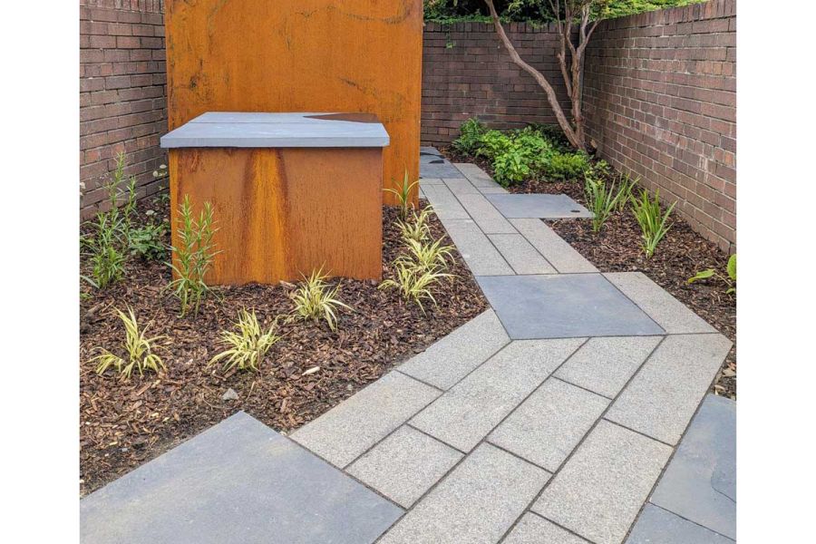 Angled path of dark grey granite planks with square blue pavers runs between rusted steel storage containers and wall.