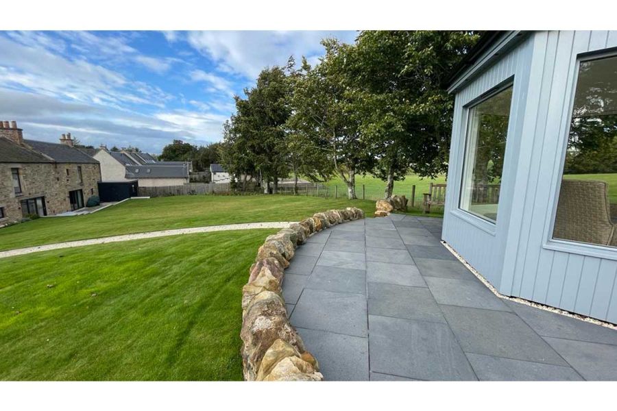 Dry stone wall creates curved edge to Midnight Black limestone patio with studio that sits above lawn sloping down to house.