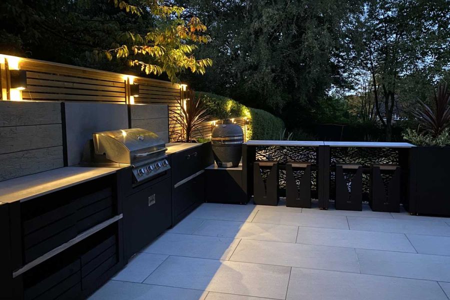 Night view of lit-up outdoor kitchen, counters on 2 sides, paved Urban Grey 900x600 porcelain paving. Design by Creative Gardens.