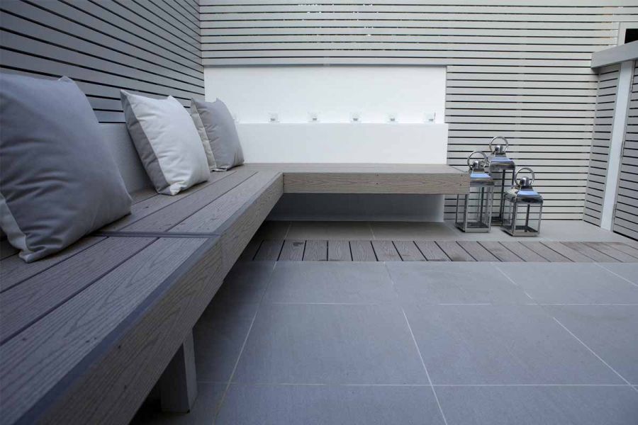 Long bench in composite decking, seen from end, against grey-painted batten fencing, with Urban Grey porcelain paving.