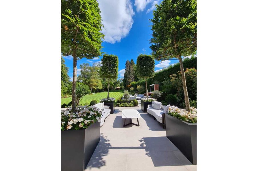 2 white matching sofas on either side of coffee table on Urban Grey Porcelain Paving in garden with trees in trough planters.