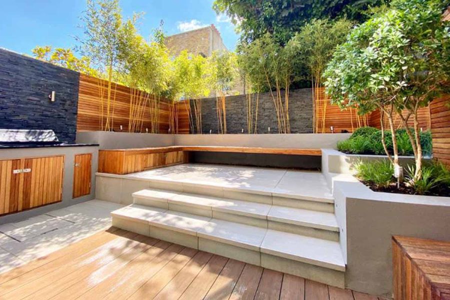 Urban Grey Porcelain steps up to paved area. A wooden bench lines raised beds filled with trees. Fence and stone cladding behind.