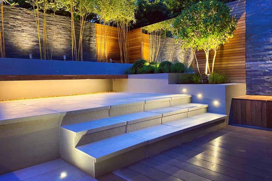 Urban Grey Porcelain steps with 40mm downstand rise from decking to paved area edged by trees lit by uplighters in raised beds.