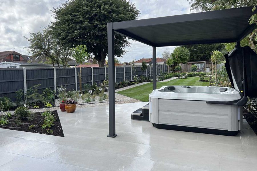 Wet weather shows off Urban Grey Porcelain Paving in large garden surrounded by black fencing. Hot tub sits underneath metal pergola.