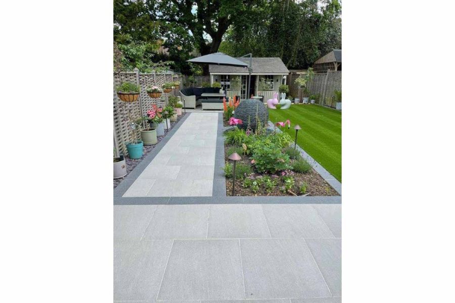 Path paved in Urban grey porcelain paving, edged in dark grey, next to long border leading to seating area covered by parasol.
