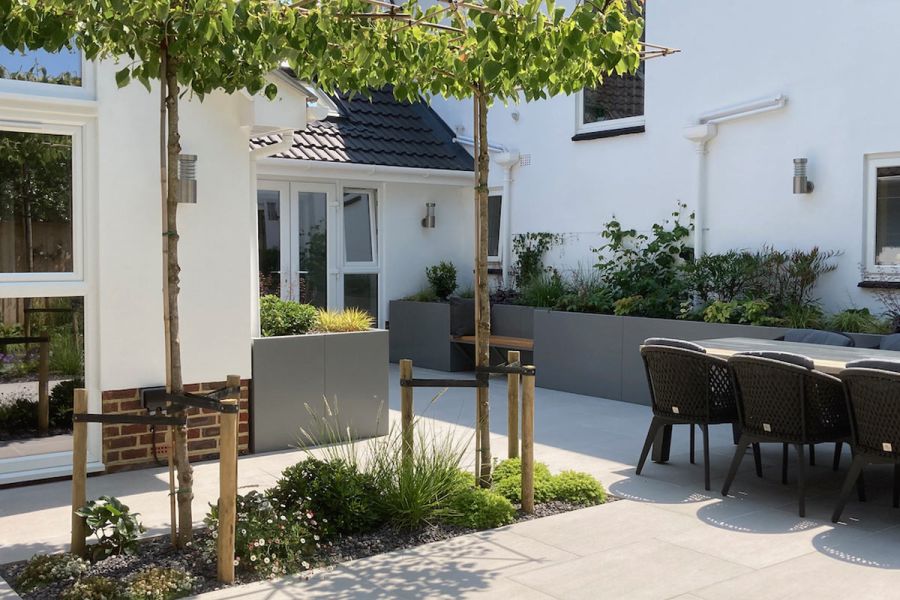 Back garden of white house paved in Urban Grey Porcelain with grey rendered raised beds, boxhead trees and garden furniture.