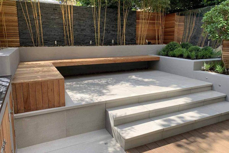 3 steps up to raised area paved in Urban Grey Porcelain tiles. Terraced beds to right. Cedar fence panels alternate with cladding.