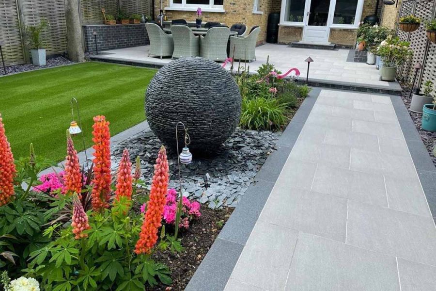 Flower bed with lupins and globe water feature edges urban grey porcelain path leading to patio with dining set at back of house.