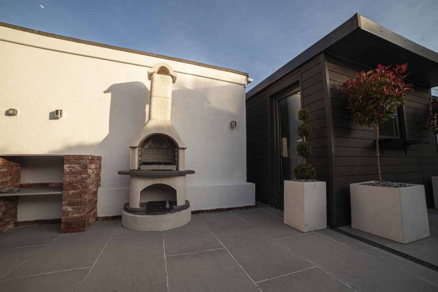 Against a white wall a white chiminea stands on urban grey vitrified porcelain paving. White planters outside garden room on right.