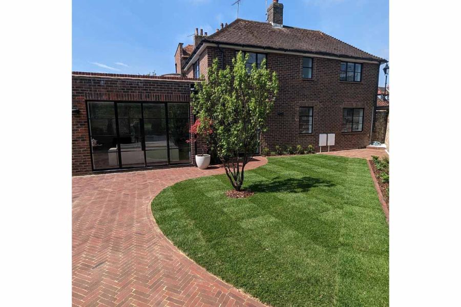 Asymmetric curve-edged lawn with small tree bordered by wide area of Abbey Dark Multi clay pavers in front of brick house.