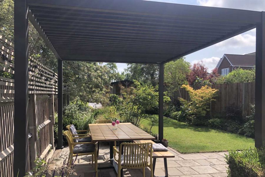 Lawn and trees in long back garden seen from under Proteus Grey aluminium pergola with louvred roof on patio of natural paving.