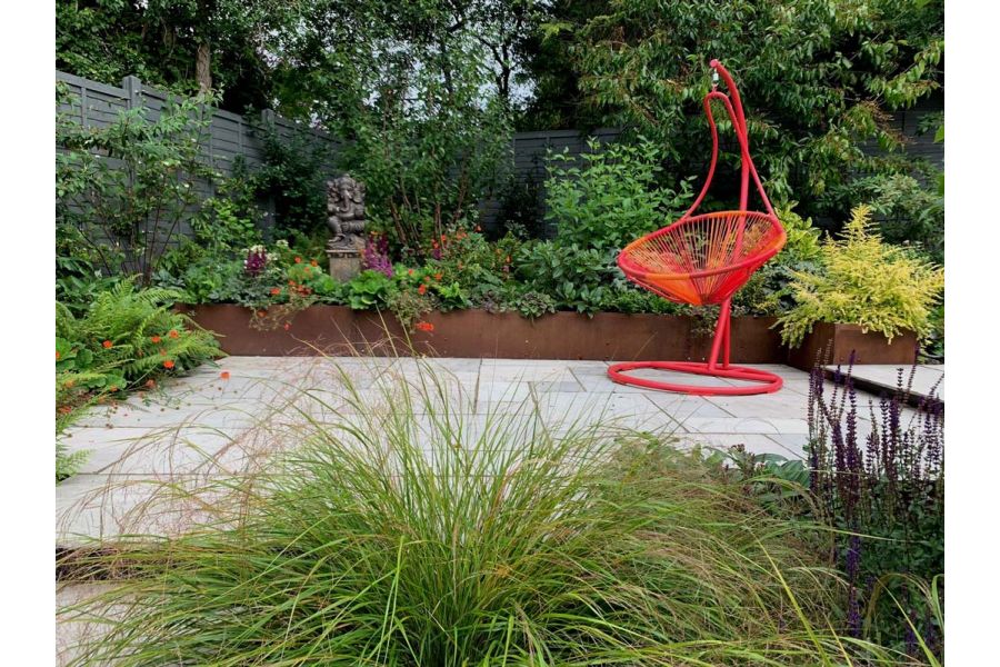 Hanging chair on rectangular patio of Kandla Grey tumbled Indian sandstone slabs, with raised beds of Corten steel lushly planted.