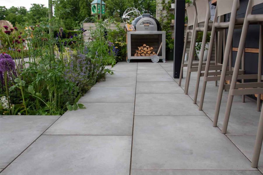 Near-ground view trade stand laid with Venetian Grey porcelain vitrified paving, with planted bed, legs of bar set and pizza oven.