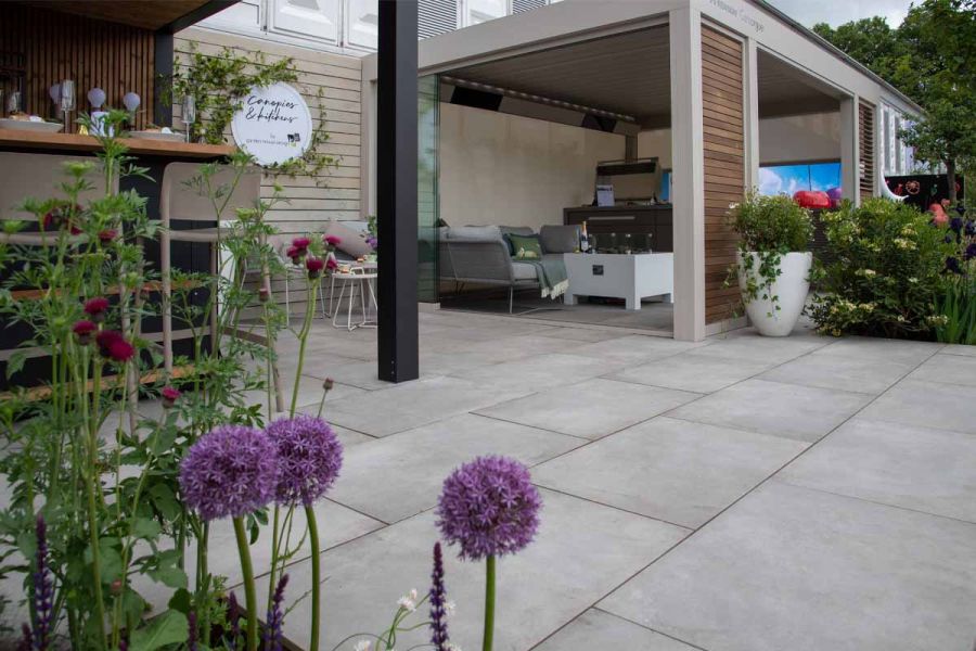 RHS Chelsea trade stand with metal pergola and furniture, planters, bar set and alliums, laid with Venetian Grey Porcelain Paving.