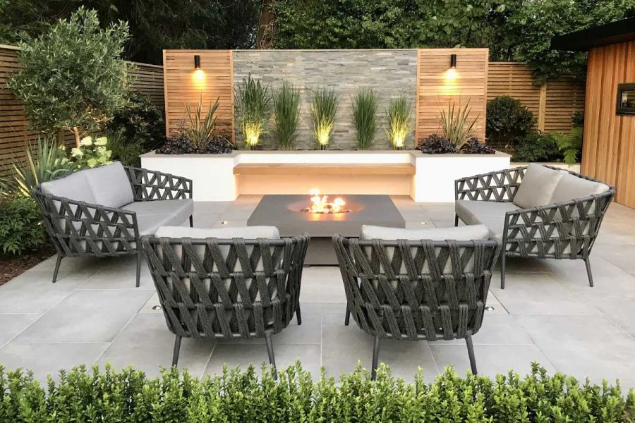 Venetian Grey Porcelain Paving with grey firepit in the middle, with 4 rattan armchairs surrounding, downlit fence in the background.