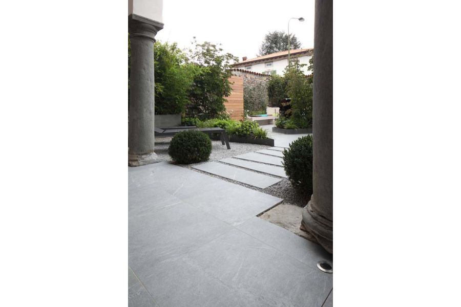 Versilia Porcelain Paving flanked by Doric pillars, giving onto plank paving set in gravel, paved area, beds and shrubs beyond.