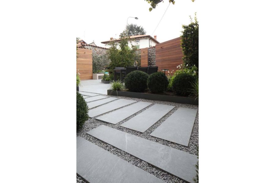 Widely spaced Versilia Porcelain patio tiles, laid into toning grey gravel, edged by topiary balls, tall fence and wall behind.