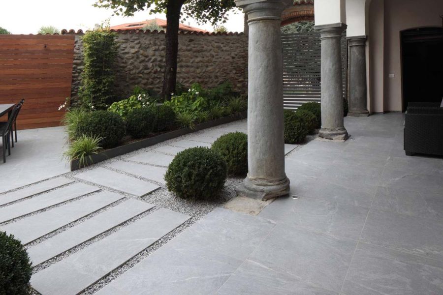 View from loggia. Stone columns match grey Versilia Porcelain paving slabs edging triangular bed with topiary, tree and shrubs.