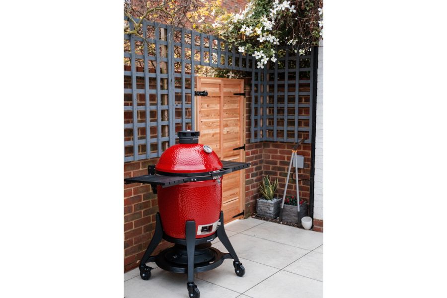 Red cooking oven in the corner of small back garden sitting on top of a porcelain patio laid in a rectangle grid laying pattern.